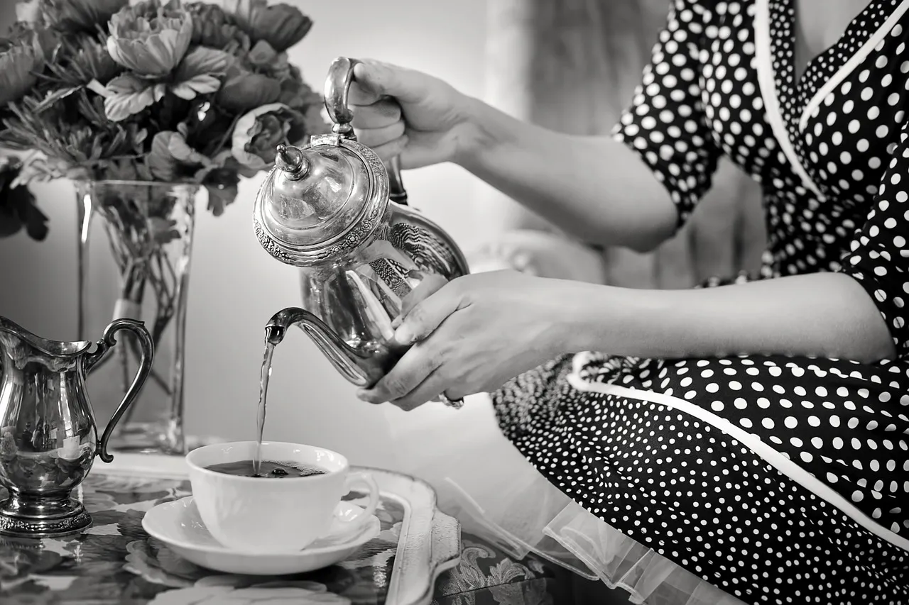 A black and white photo of tea being poured from a silver teapot at a table