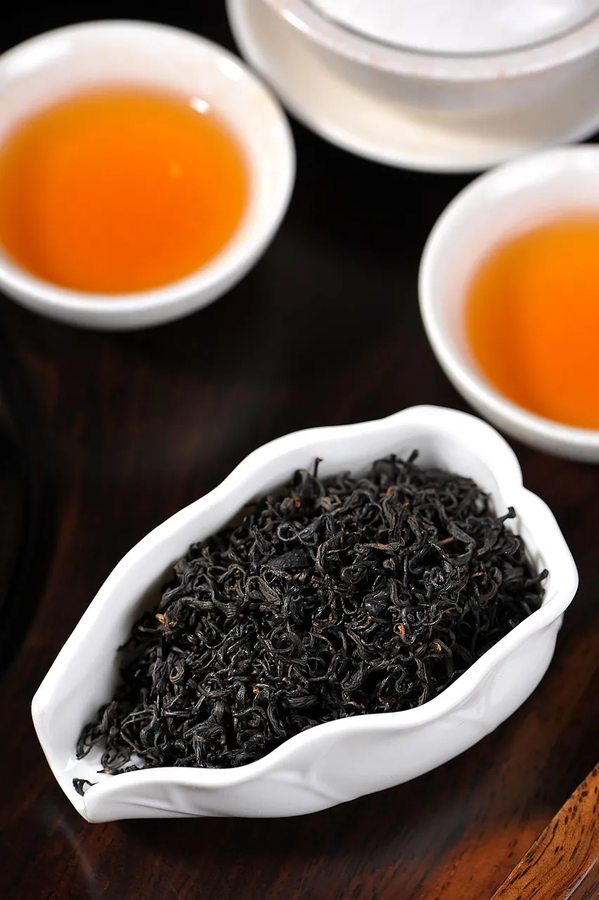 Twisted black tea leaves in a bowl next to cups of amber-red tea