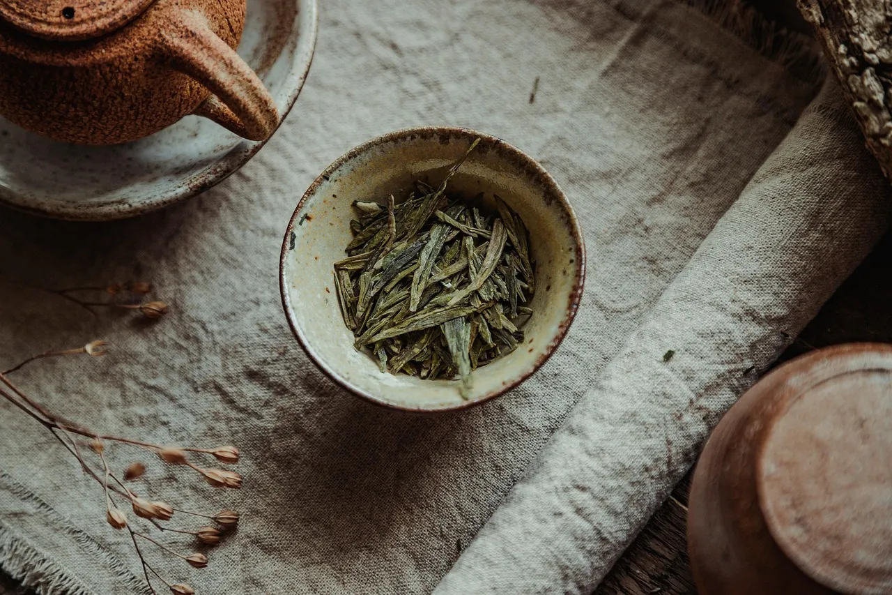 Flat green Longjing tea leaves in a small bowl beside a teapot