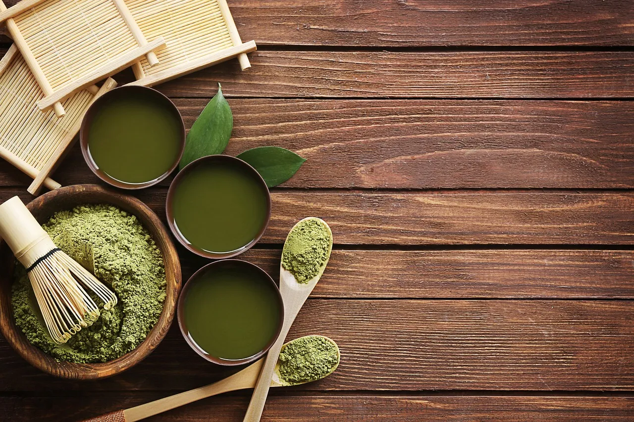 Matcha powder, a bamboo whisk and cups of whisked matcha on a wooden table
