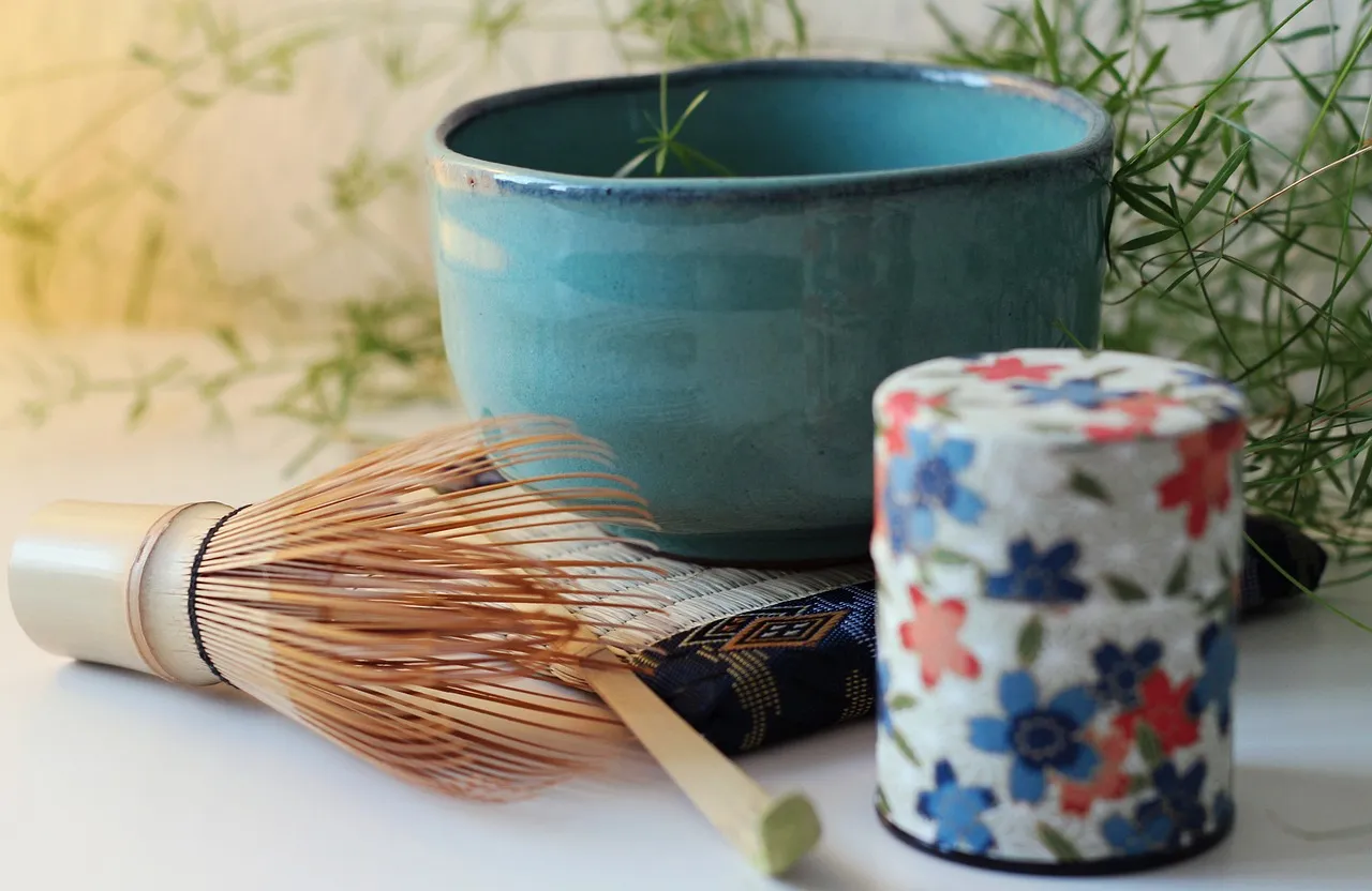 A bamboo whisk beside a turquoise tea bowl and a patterned tea caddy