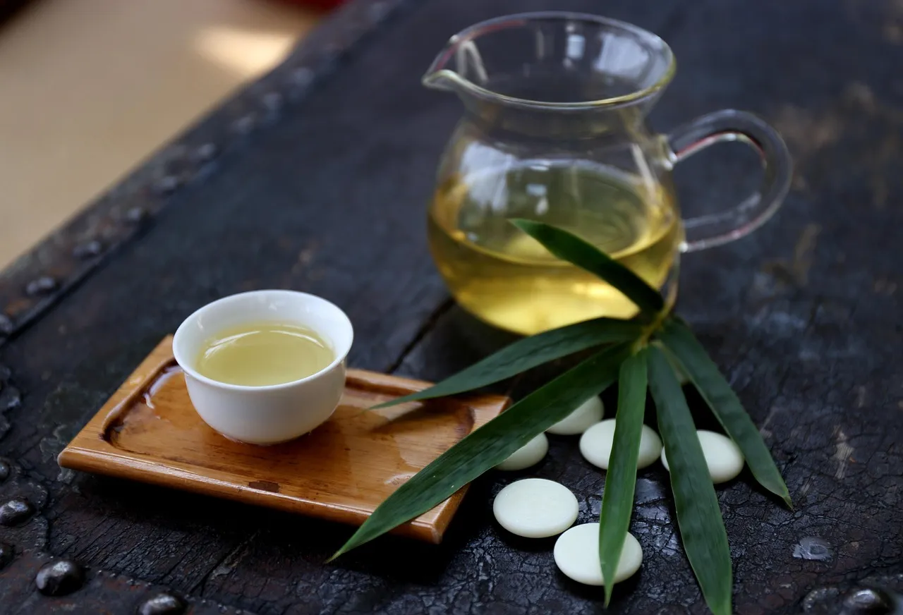 A glass pitcher of pale tea and a small tasting cup on a dark wooden tray